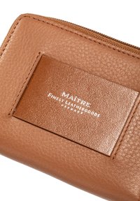 Hellbraune Lederbörse mit strukturierter Oberfläche, versehen mit einem glatten rechteckigen Patch mit weißer Prägung. Reißverschluss entlang der oberen Kante.