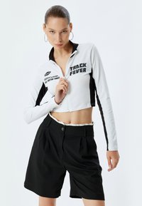 Giacca cropped bianca con zip e dettagli neri, decorata con la grafica "TRACK FEVER", abbinata a pantaloni corti neri a vita alta con pieghe frontali.