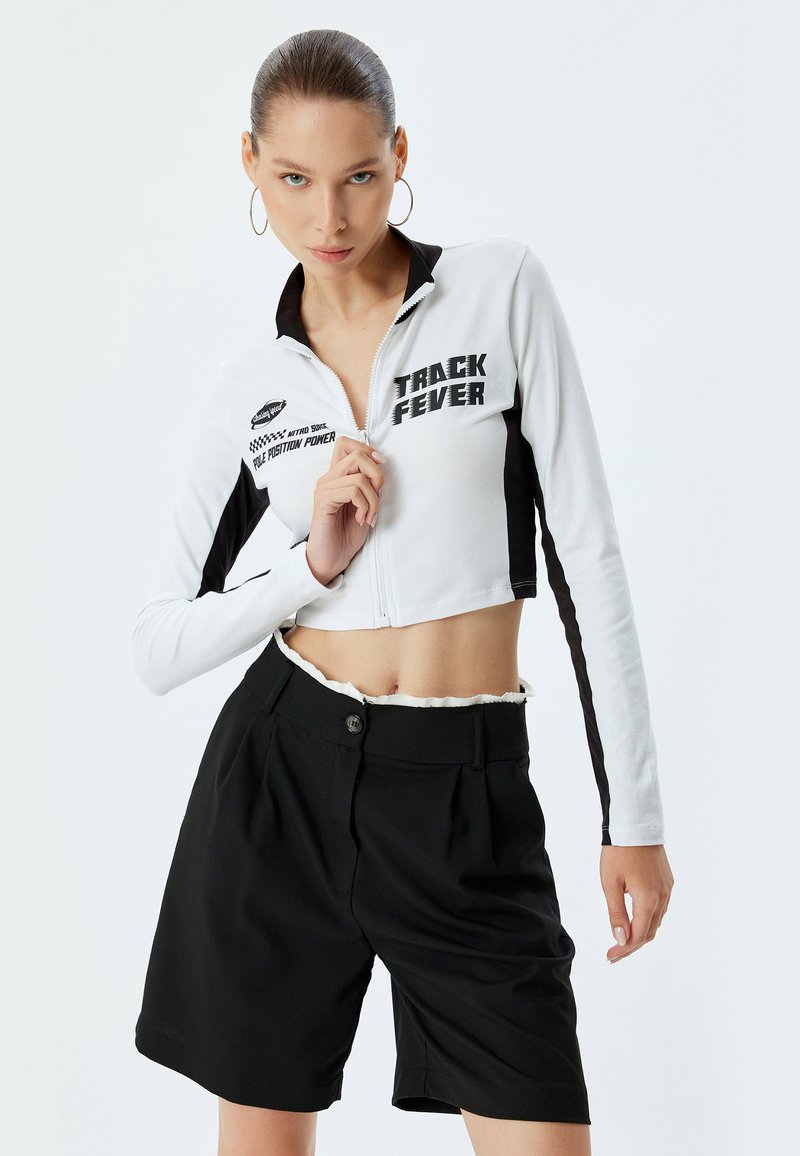 Giacca cropped bianca con zip e dettagli neri, decorata con la grafica "TRACK FEVER", abbinata a pantaloni corti neri a vita alta con pieghe frontali.