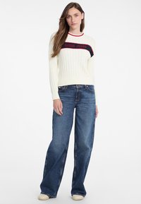 Pulover crem cu dungi, având un logo "GUESS JEANS" roșu și negru, asociat cu blugi wide-leg de culoare albastră închisă și adidași beji, pe un fundal simplu.