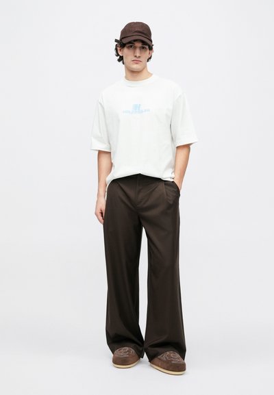 Valge üle suurusega t-särk sinise tekstiga, laiad pruunid püksid ja pruun müts logoga. Pruunid sussid lillemustrilise aktsendiga.