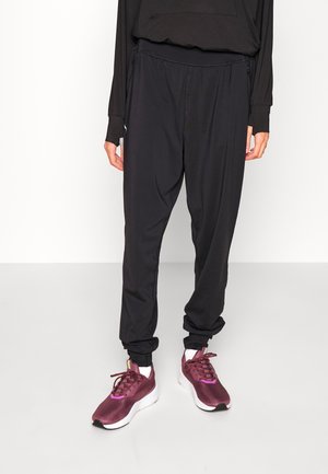 Osoba nosząca luźne czarne spodnie i bordowe trampki z białymi podeszwami, stojąca na tle jednolitej białej ściany.