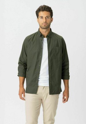 Mężczyzna stojący, ubrany w rozpiętą ciemnozieloną koszulę na białej koszulce i beżowe spodnie na jednolitym białym tle.