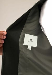 Czarna marynarka, z zielonym wykończeniem. Posiada metkę z napisem "NEXT," rozmiar "40R," oraz "WYPRODUKOWANO W Wietnamie." Gładka tekstura, strukturalny design.