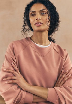 Frau mit lockigem Haar, die einen rosa Sweatshirt trägt, mit verschränkten Armen und leicht zur Seite blickend, vor einem schlichten beigen Hintergrund.