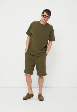 Mand iført olivengrøn oversized t-shirt og matchende shorts med brune slip-on sko, stående med hænderne i lommerne mod en hvid væg.