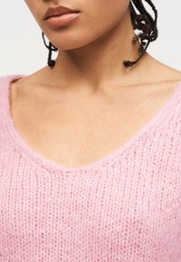 Gros plan sur une personne portant un pull en maille rose doux à col en V, montrant les lèvres, le cou, les clavicules et des mèches de cheveux tressés.
