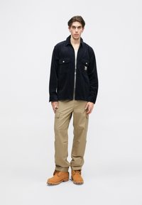 Marinblå kordflöjels zipjacka med två bröstfickor, kombinerad med beige cargobyxor och bruna arbetsskor, har en avslappnad passform och strukturerad textil.