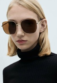 Lunettes de soleil surdimensionnées teintées de brun avec une monture en métal doré, présentant un design géométrique. Le modèle porte un haut en col roulé côtelé noir.