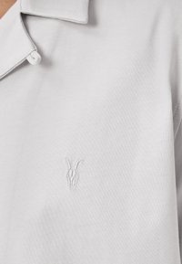 Lys grå polo shirt lavet af tekstureret stof, med krave og et subtilt broderet logo på venstre side.