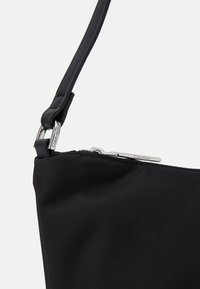 Sac à main en nylon noir avec une texture lisse, doté d'une fermeture éclair argentée et d'une étiquette en métal de marque. La bandoulière est réglable et élégante.