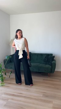 Top bianco senza maniche con dettaglio annodato sul davanti, abbinato a pantaloni neri a vita alta e a gamba larga. Divano verde e pavimento in legno sullo sfondo.