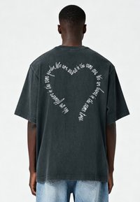 Tumšpelēka, pārāk liela kokvilnas t-kreklu ar izliektiem baltajiem tekstiem sirds formā uz muguras, īsām piedurknēm un brīvu piegriezumu.