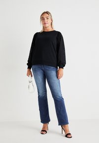 Black sweatshirt with textured, puffed sleeves, paired with blue flared jeans and black heeled sandals. Holds a white handbag.