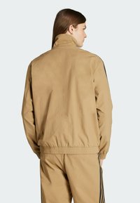 Chaqueta ligera de color marrón claro con cuello alto y puños elásticos. Presenta rayas oscuras a lo largo de los lados y un diseño de corte relajado.
