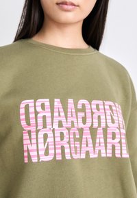 Olivovo zelená mikina s okrúhlym výstrihom, na ktorej sú v odvážnom, pruhovanom ružovom písme napísané slová "NØRGAARD" a "RECYCLED" v nadrozmernom dizajne. Mäkko sa nosí.