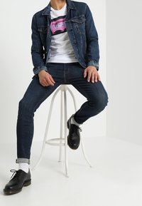 Chaqueta vaquera con botones de metal, camiseta blanca con gráfico y texto "LEVI'S", vaqueros oscuros y zapatos negros con cordones. Sentado en un taburete blanco.