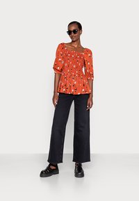 Top floral vermelho com mangas bufantes e corpete franzido, combinado com jeans pretos de perna larga e sandálias pretas chunky. Mulher a usar óculos de sol.