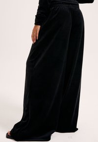 Zwarte wijde broek van zachte veloursstof, met een elastische tailleband en subtiele textuurdetails langs de zijkanten.