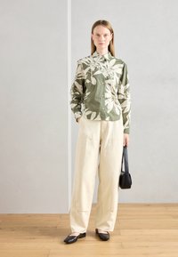 Chemise à boutons verte à motif floral avec des motifs blancs ; pantalon large beige ; ballerines noires ; petit sac à bandoulière noir ; sol en bois en arrière-plan.