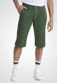 Șorturi cargo din bumbac verde, cu un design ajustat, dotate cu buzunare și o tăietură până la genunchi, combinate cu șosete albe și adidași.