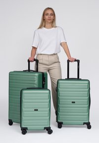 Tre valigie rigide verdi con design a rilievo lineare, finitura opaca e maniglie retrattili, posizionate l'una accanto all'altra.