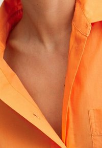 Chemise orange vif avec col et poche poitrine, en coton lisse avec une coupe légèrement décontractée, mettant en valeur une couleur vibrante et des détails de design.