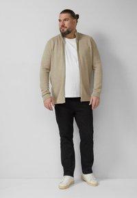 Beżowy kardigan na zamek na biały t-shirt, w połączeniu z czarnymi spodniami i białymi trampkami, na neutralnym tle.