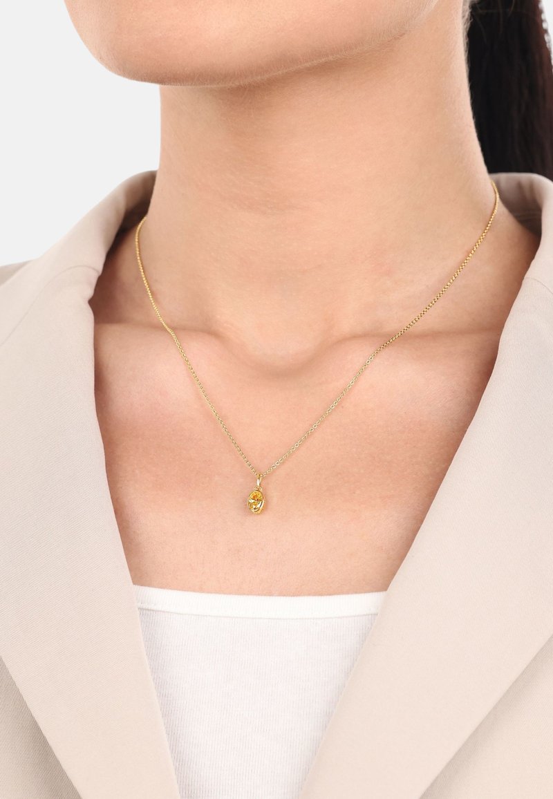 Collana d'oro con una catena sottile e un piccolo pendente ovale con un gemma gialla, indossata su una maglietta bianca, su uno sfondo neutro.