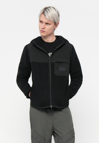 Sort fleece zip-up hoodie med hætte, kontrasterende øverste sektion, frontlomme og diskret logo-detail på brystet. Tekstileret stof.