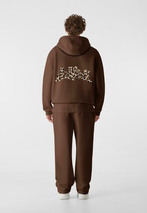 Brun hoodie och matchande sweatpants i mjukt material. Hoodien har en stor leopardmönstrad logotyp på ryggen. Vita sneakers synliga.