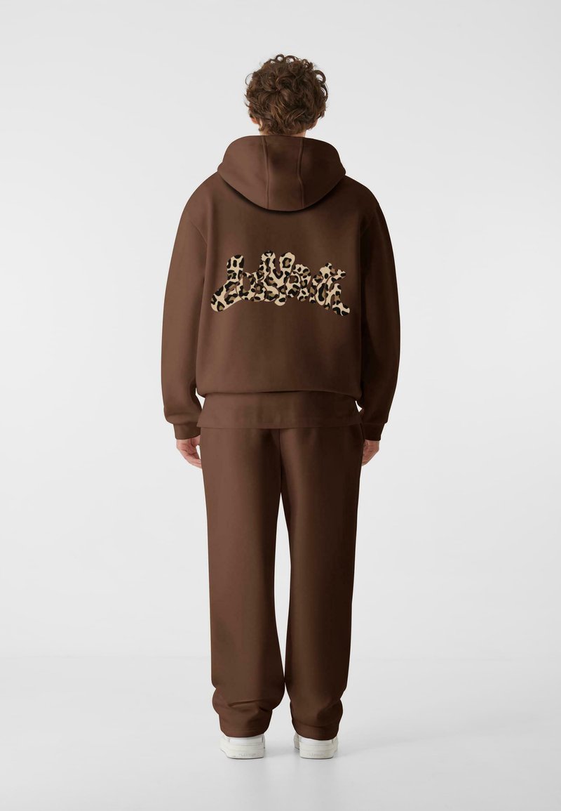 Brun hettegenser og matchende joggebukser laget av mykt materiale. Hettegenseren har et stort, leopardmønstret logo på ryggen. Hvite sneakers er synlige.