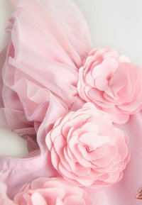 Vaaleanpunainen kangas, jossa kaula-aukon kohdalla kerroksittain verkkoa ja teksturoituja keinokukkia, pehmeine terälehtineen ja kerättyine yksityiskohtineen.