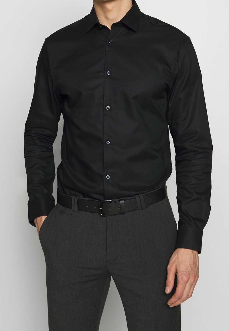Camicia nera a maniche lunghe con colletto a punta, dotata di bottoni blu e una leggera lucentezza, abbinata a pantaloni grigi e una cintura nera.