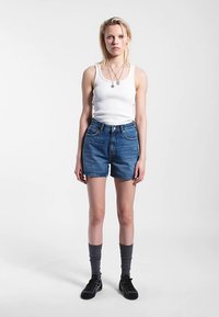 Modré džínové šortky s vysokým pasem, bílý tílko, šedé podkolenky a černé boty. Model stojí před jednoduchým bílým pozadím.