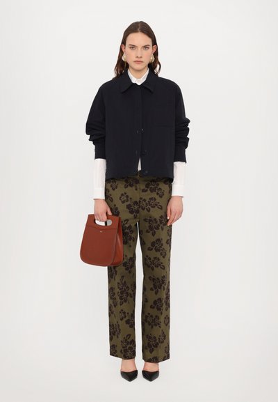 Zwarte button-up jas over een witte shirt, olijfgroene broek met zwart bloemenpatroon, bruine handtas en zwarte puntige schoenen.