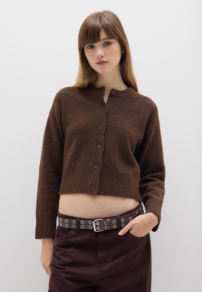 Cárdigan marrón cortado con escote redondo, cierre de botones y dobladillo acanalado. Combinado con pantalones marrones oscuros que presentan detalles de cinturón con ojales metálicos.