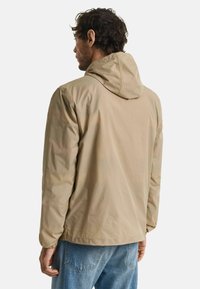 Giacca khaki con cappuccio, dalla texture liscia, caratterizzata da un design semplice e polsini elastici, indossata sopra jeans blu.