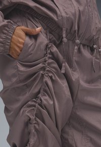 Combinaison en tissu grise froncée avec des poignets élastiques, fermeture éclair et cordons ajustables. Présente une poche intégrée dans le design.