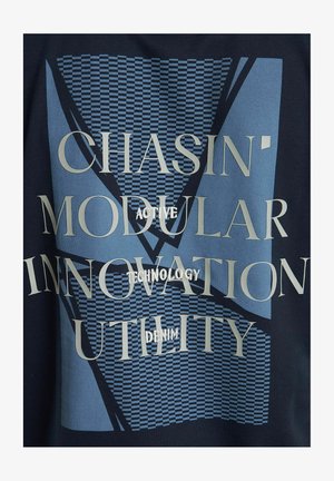 Sotilaslaivaston sininen kangas, jossa on geometrisia kuvioita ja suurta tekstiä kullassa ja vaaleansinisessä. Korostaa sanoja kuten "Chasin'" ja "Modular Technology".