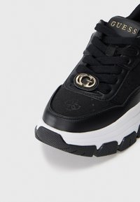 Sneaker nera con tomaia in tessuto testurizzato, suola in gomma, intersuola bianca e dettaglio del logo dorato sulla linguetta. Accenti di cuciture distintive.