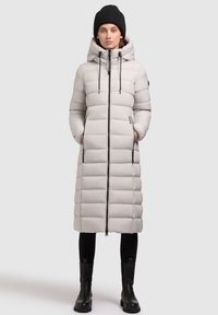 Lichtgrijze pufferjas met een capuchon en zwarte accenten, voorzien van een rits aan de voorkant, verticale bies en zijzakken. Draagt met zwarte leggings en laarzen.