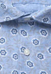 Chemise bleu clair avec petits motifs floraux bleus et blancs, col plié et bouton blanc visible près du centre.