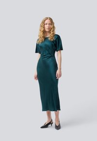 Femme aux cheveux bouclés blonds, portant une robe midi en satin teal et des chaussures à talons kitten noires à bride arrière, se tenant devant un fond gris clair.