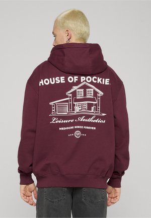 Lühikeste blondide juustega inimene kannab tumepunast kapuutsiga dressipluusi, mille seljal on maja visand ja tekst "HOUSE OF POCKIES Leisure Aesthetics Mediocre Since Forever".