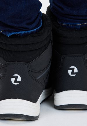 Zwarte hoge sneakers met een gestructureerde afwerking, witte accenten, een gewatteerde kraag en een logo op de hiel, neergezet tegen denim jeans.