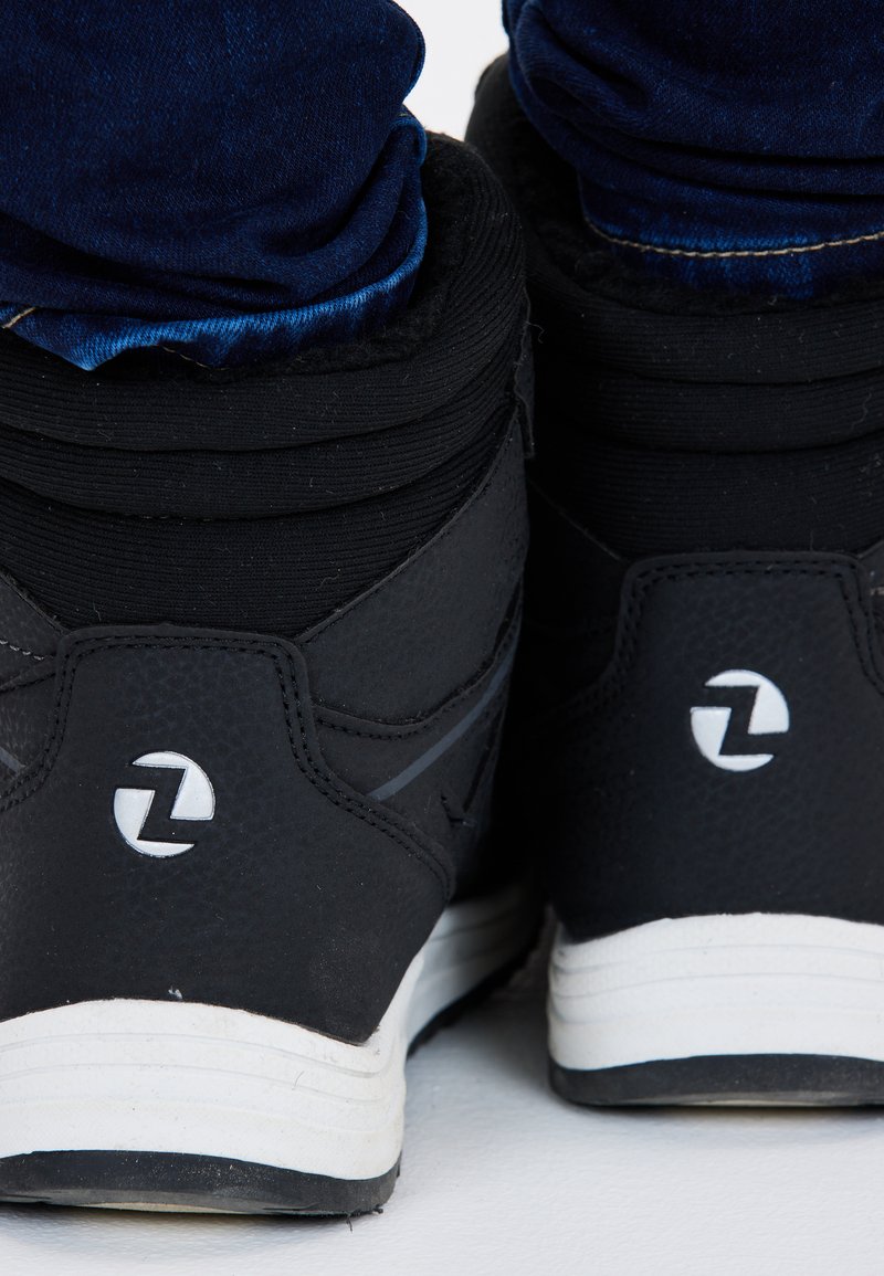 Sorte høj-top sneakers med en tekstureret overflade, hvide accenter, polstret krave og et logo på hælen, sat mod denimjeans.
