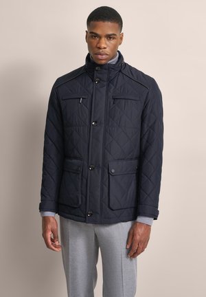 Mann trägt eine marineblaue gesteppte Jacke mit geknöpften Taschen und hellgrauen Hosen vor einem schlichten Hintergrund.