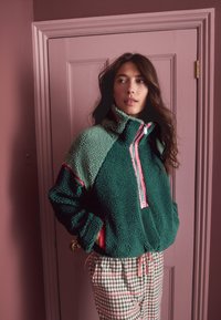 Roheline fliispusa kõrge kraega, roosa tõmblukuga ja värvilise plokkkujundusega. Kandmisel ruudulised püksid, millel on rohevalged ruudud.