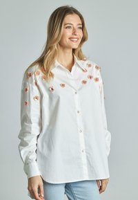 Camicia bianca con ricami floreali e paillettes sulle spalle, con maniche lunghe e orlo curvo.